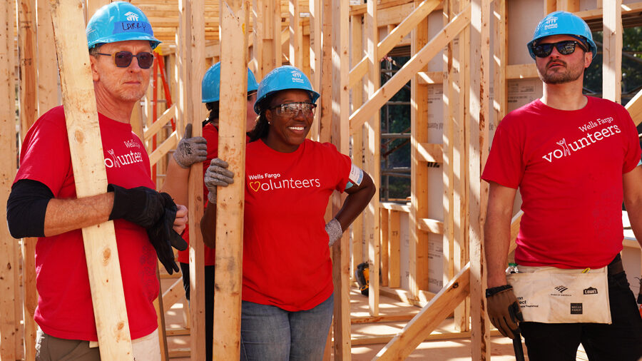 Four people in red volunteer shirts and blue construction hats stand in the wooden frame of a home.