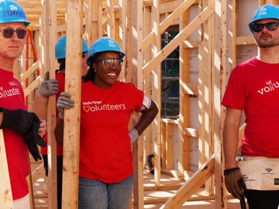 Four people in red volunteer shirts and blue construction hats stand in the wooden frame of a home.