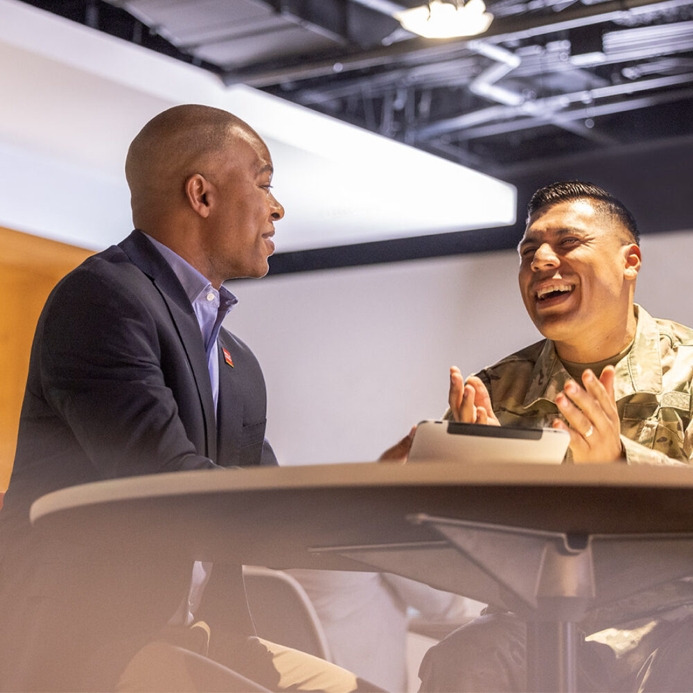 Two people — one in a business suit and the other in military camouflage — sit at a table and talk.