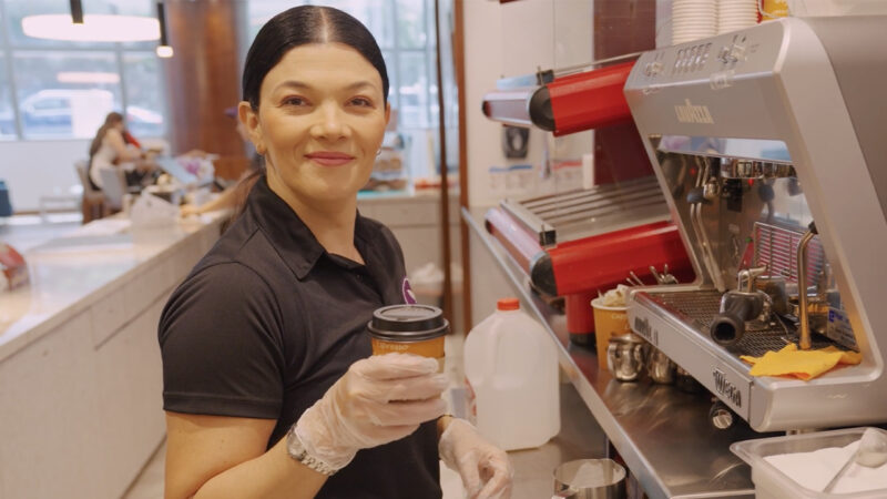 A woman in a smoothie bar is holding a cup with a lid and smiling.