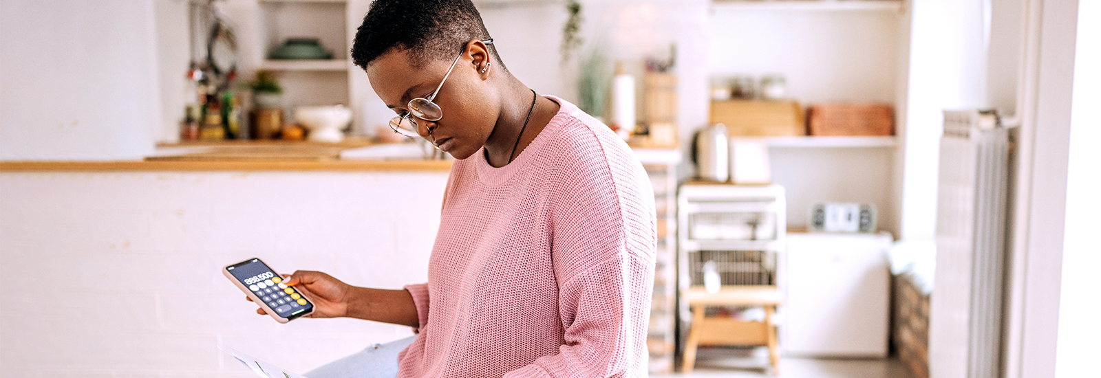 A person sits in a kitchen holding a smart phone open to a calculator in one hand and a bill in the other hand.