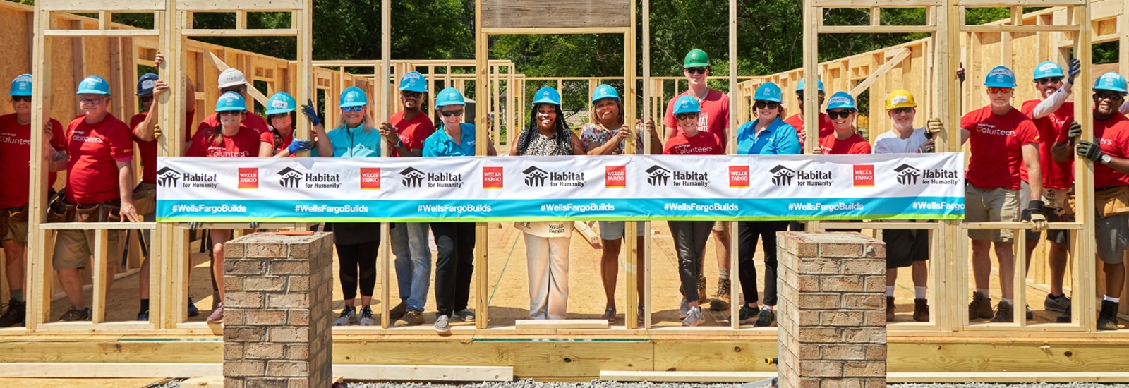 A group of people in hardhats stand inside the frame of a partially built home. A Wells Fargo and Habitat for Humanity banner is in front of them.