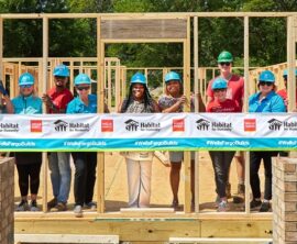 A group of people in hardhats stand inside the frame of a partially built home. A Wells Fargo and Habitat for Humanity banner is in front of them.