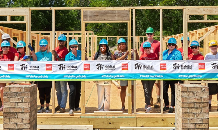 A group of people in hardhats stand inside the frame of a partially built home. A Wells Fargo and Habitat for Humanity banner is in front of them.