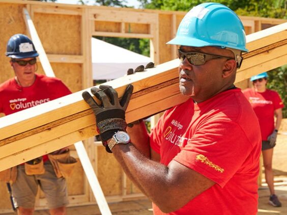 A man in a hardhat carries wood planks at a construction site.