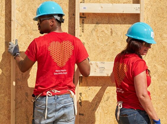 Two people in hardhats stand in front of the wall of a partially built home.