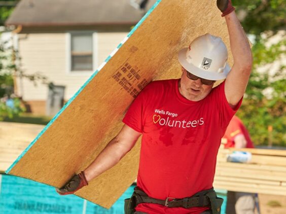 A person in a hardhat carries a plywood panel at a construction site.