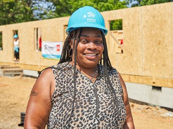 A woman in a hardhat smiles as she stands in front of a partially built home.