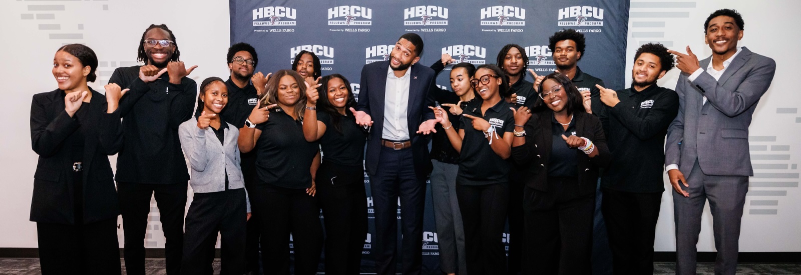 A dozen or so smiling college students point toward a professional football player in the center of the group.