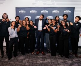 A dozen or so smiling college students point toward a professional football player in the center of the group.