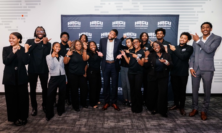 A dozen or so smiling college students point toward a professional football player in the center of the group.