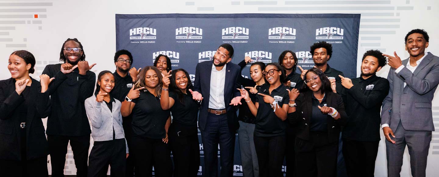 A dozen or so smiling college students point toward a professional football player in the center of the group.