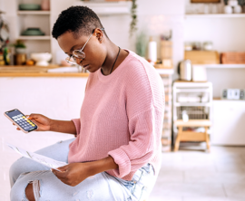 A person sits in a kitchen holding a smart phone open to a calculator in one hand and a bill in the other hand.