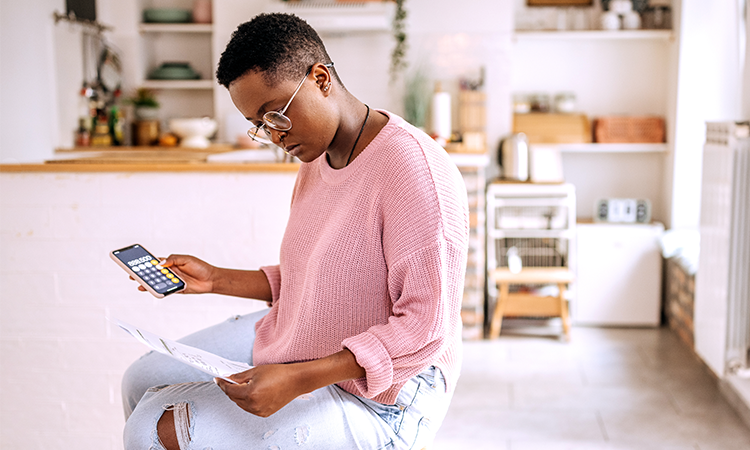 A person sits in a kitchen holding a smart phone open to a calculator in one hand and a bill in the other hand.