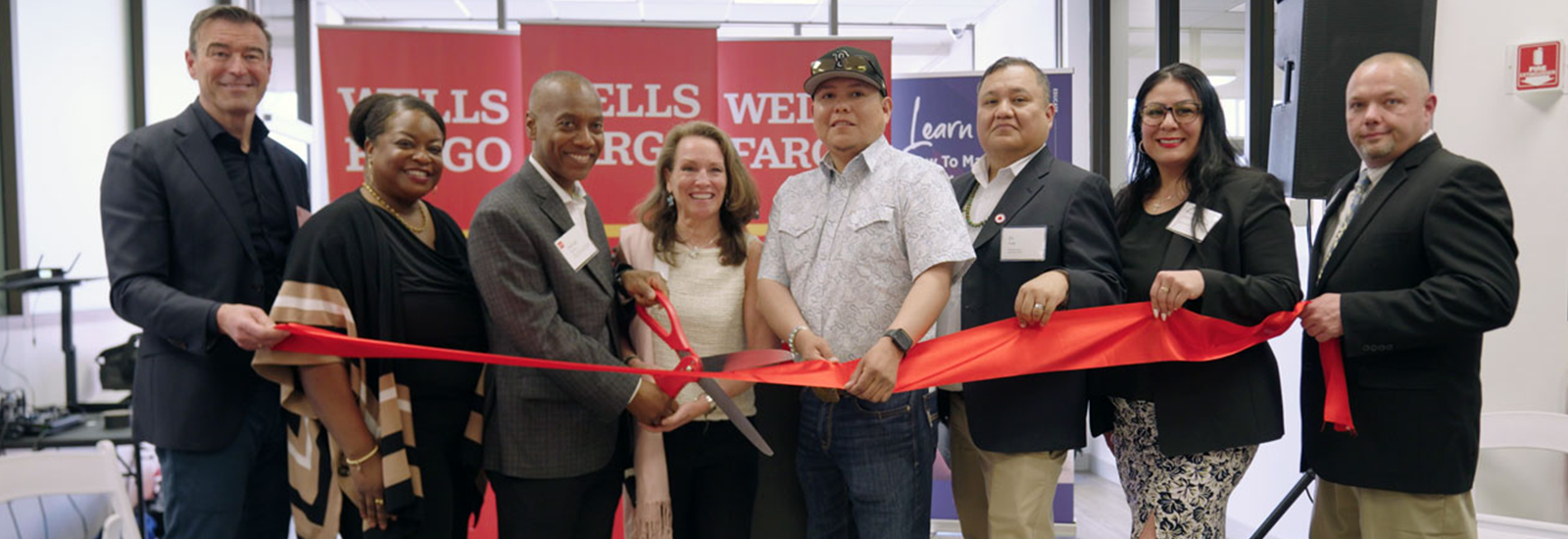 Several people stand behind a ribbon as the person near the center cuts the ribbon. 