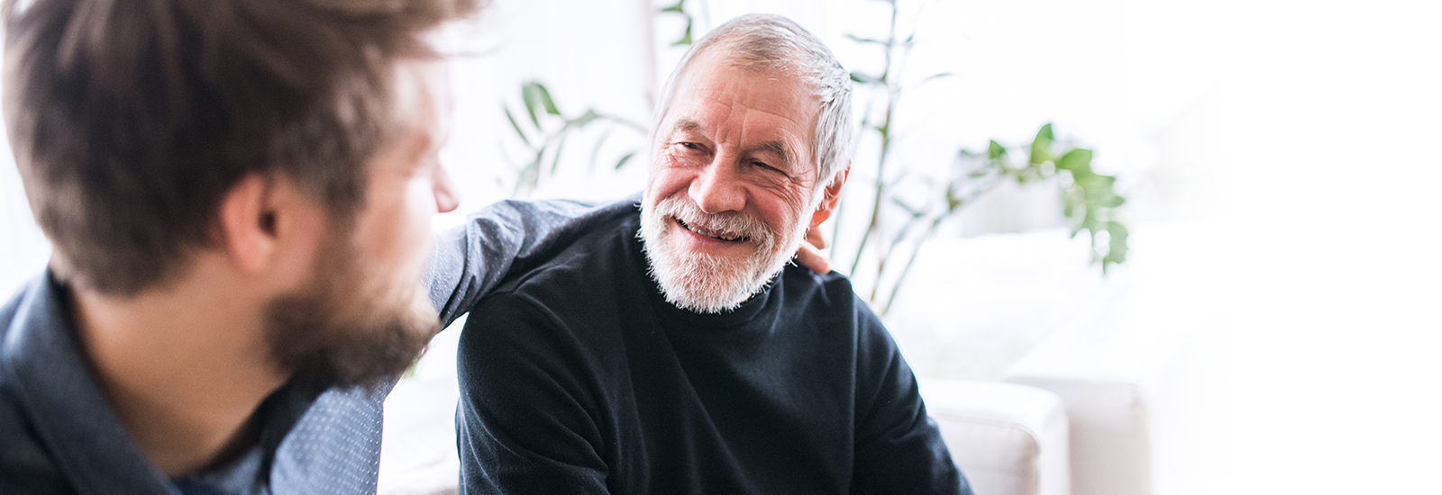 An older adult sits on the couch smiling. A younger adult sits next to him with his arm around him.