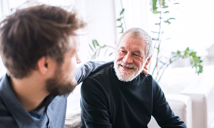An older adult sits on the couch smiling. A younger adult sits next to him with his arm around him.