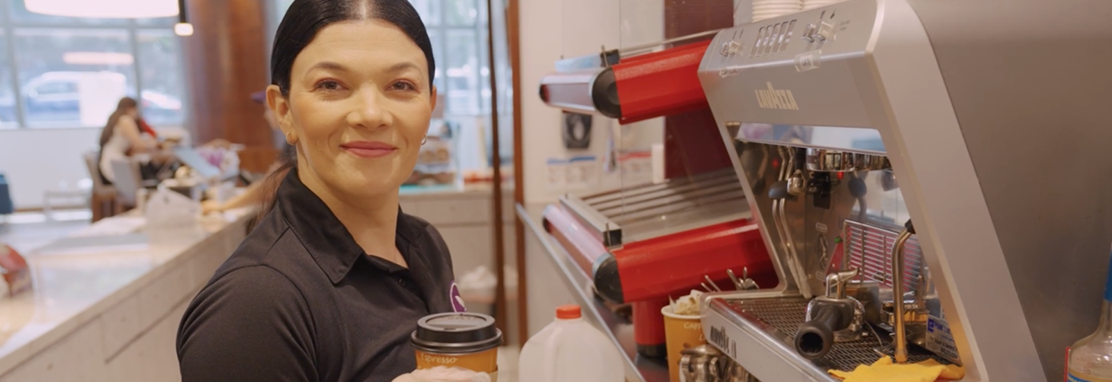 A woman in a smoothie bar is holding a cup with a lid and smiling.