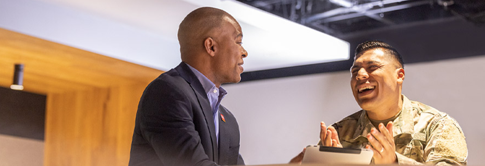 Two people — one in a business suit and the other in military camouflage — sit at a table and talk.