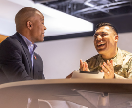 Two people — one in a business suit and the other in military camouflage — sit at a table and talk.