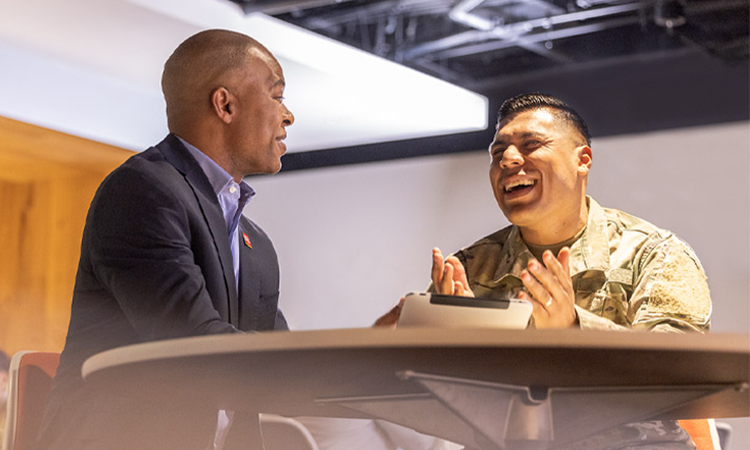 Two people — one in a business suit and the other in military camouflage — sit at a table and talk.