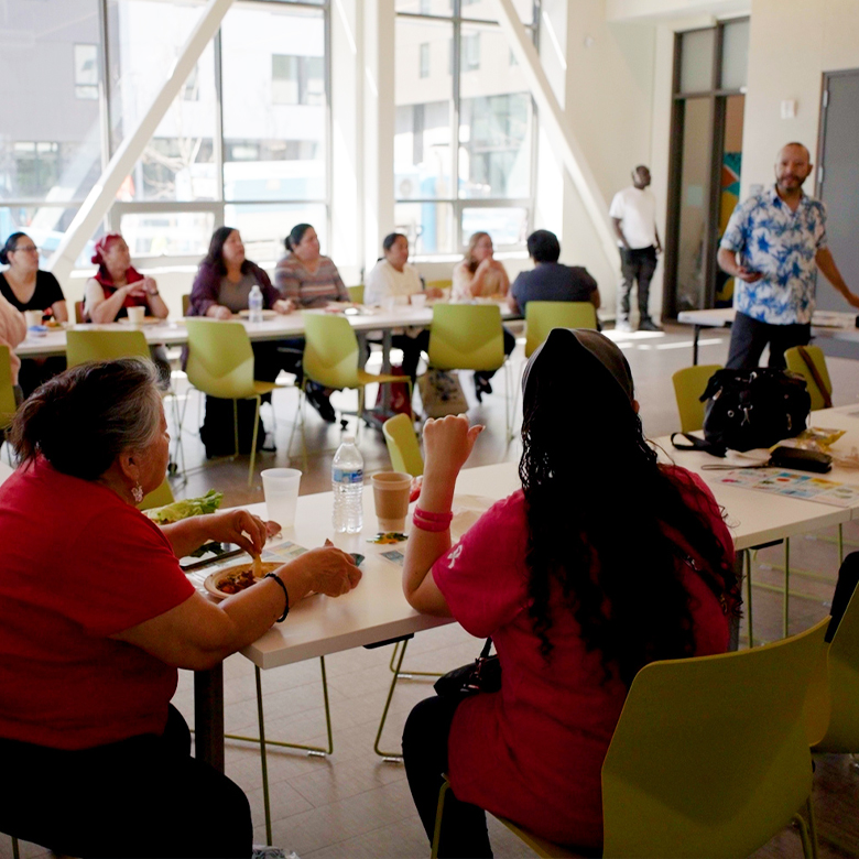 A group of people sit at a table and look at a person who is presenting at the front of the room.