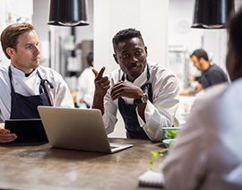 Two men wearing aprons are standing at a counter with laptops.