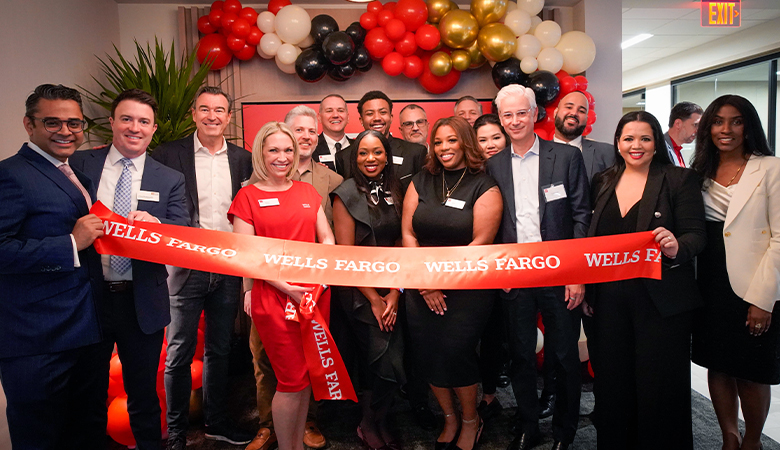 A group of people in professional attire stand behind a large red ribbon with the words Wells Fargo in white letters repeated across the ribbon