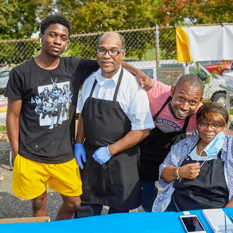 A man wearing an apron has a younger person standing next to him with his arm around him, and two other smiling people on the other side of him.
