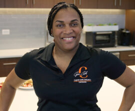 A woman in a black shirt with orange lettering stands in a kitchen
