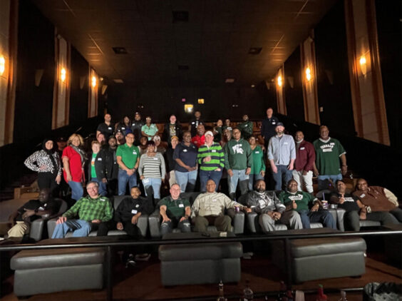 A group of people standing in a movie theater ready to watch the NCAA championship.
