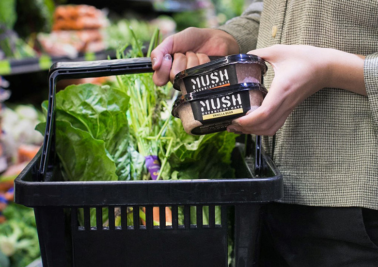 A shopper holds a shopping basket with produce and a few small tubs of MUSH overnight oats.