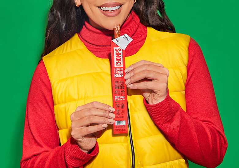 A smiling woman in a bright outfit holds a Chomps meat stick product.