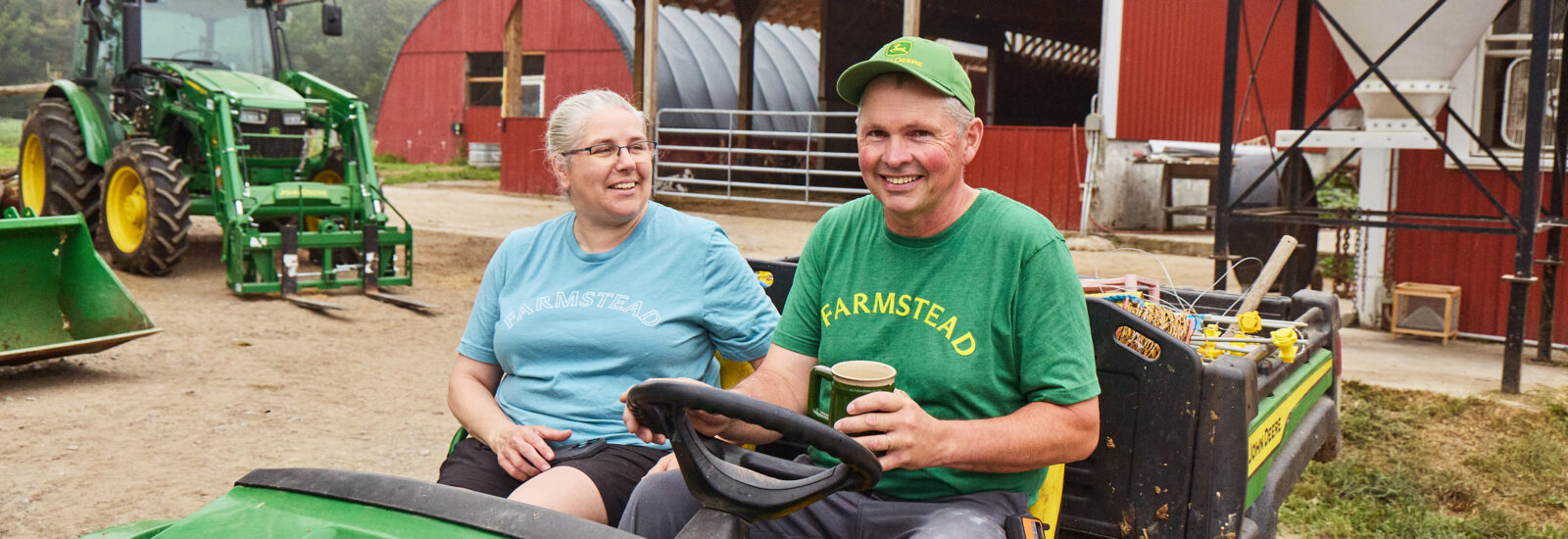 On a farm, a smiling man and a woman sit on a tractor. The man is holding a coffee cup.