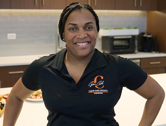 A woman in a black shirt with orange lettering stands in a kitchen