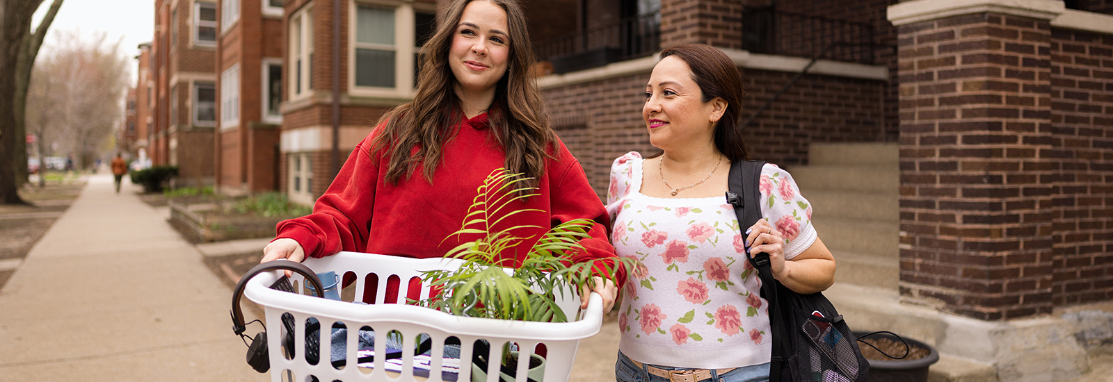 A young college student carries items for her dorm in a laundry basket as she walks alongside her mom