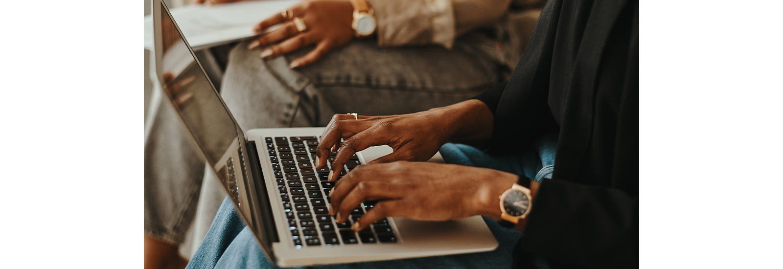 Two people, seen only from the waist down, are sitting next to each other. One is typing on a laptop and the other is holding papers.