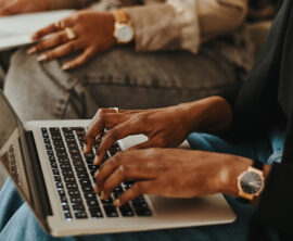 Two people, seen only from the waist down, are sitting next to each other. One is typing on a laptop and the other is holding papers.