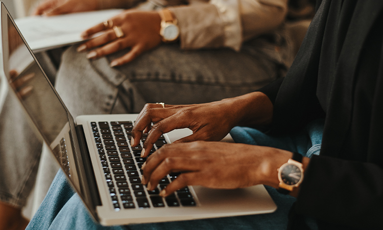 Two people, seen only from the waist down, are sitting next to each other. One is typing on a laptop and the other is holding papers.
