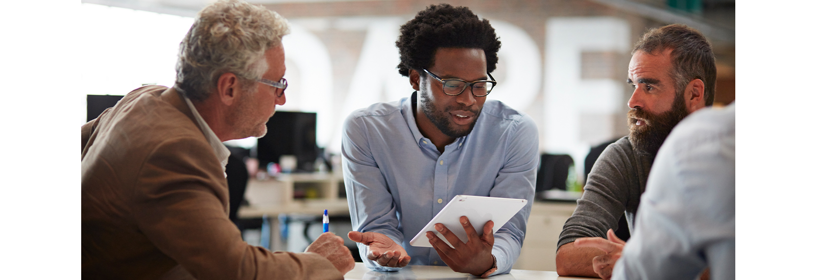 Several people sit around a table listening to the person in the center, who holds a tablet