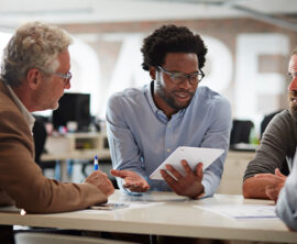 Several people sit around a table listening to the person in the center, who holds a tablet