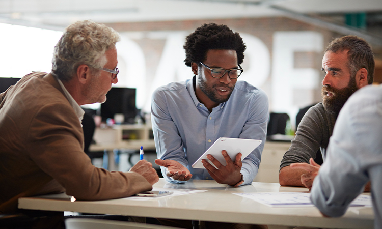 Several people sit around a table listening to the person in the center, who holds a tablet