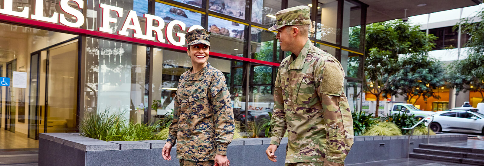 Two soldiers - a woman and a man - smile at each other while they walk past a Wells Fargo branch