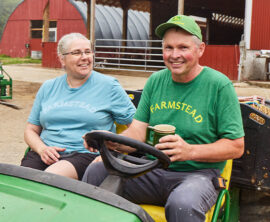 On a farm, a smiling man and a woman sit on a tractor. The man is holding a coffee cup.