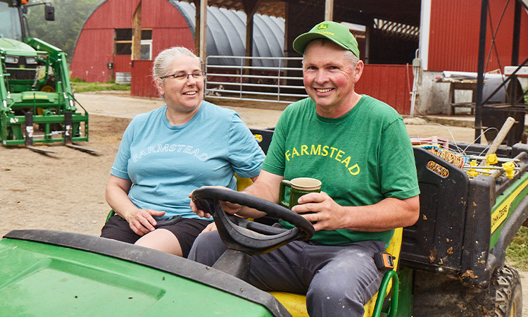 On a farm, a smiling man and a woman sit on a tractor. The man is holding a coffee cup.