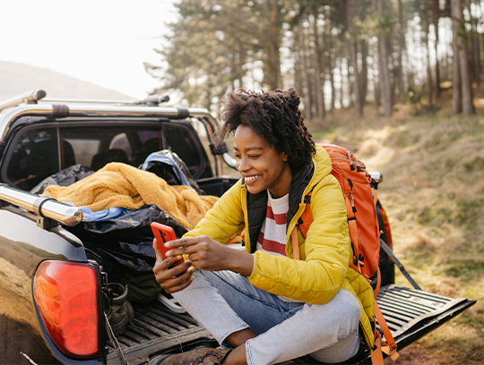 A woman in a yellow park and orange backpack sits in the open bed of a pickup truck with a forest behind her.
