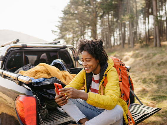 A woman in a yellow park and orange backpack sits in the open bed of a pickup truck with a forest behind her.