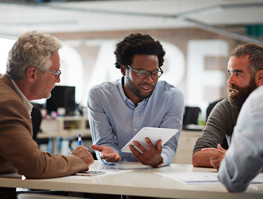 Several people sit around a table looking at a tablet.