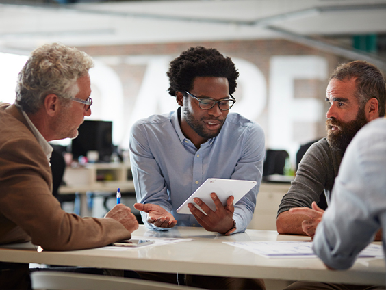 Several people sit around a table looking at a tablet.