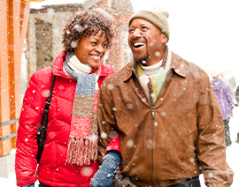 A man and a woman laugh while walking together, bundled in winter clothing, as snow falls.